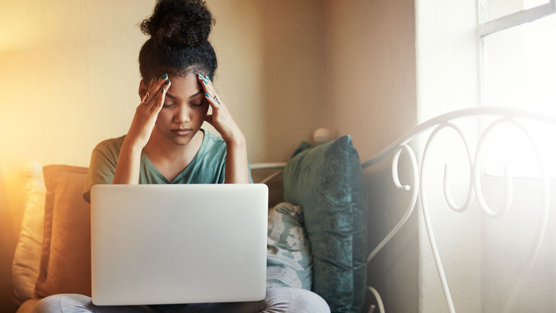 A young woman sits on her bed looking stressed with her hands resting on her temples. She has a laptop in her lap.