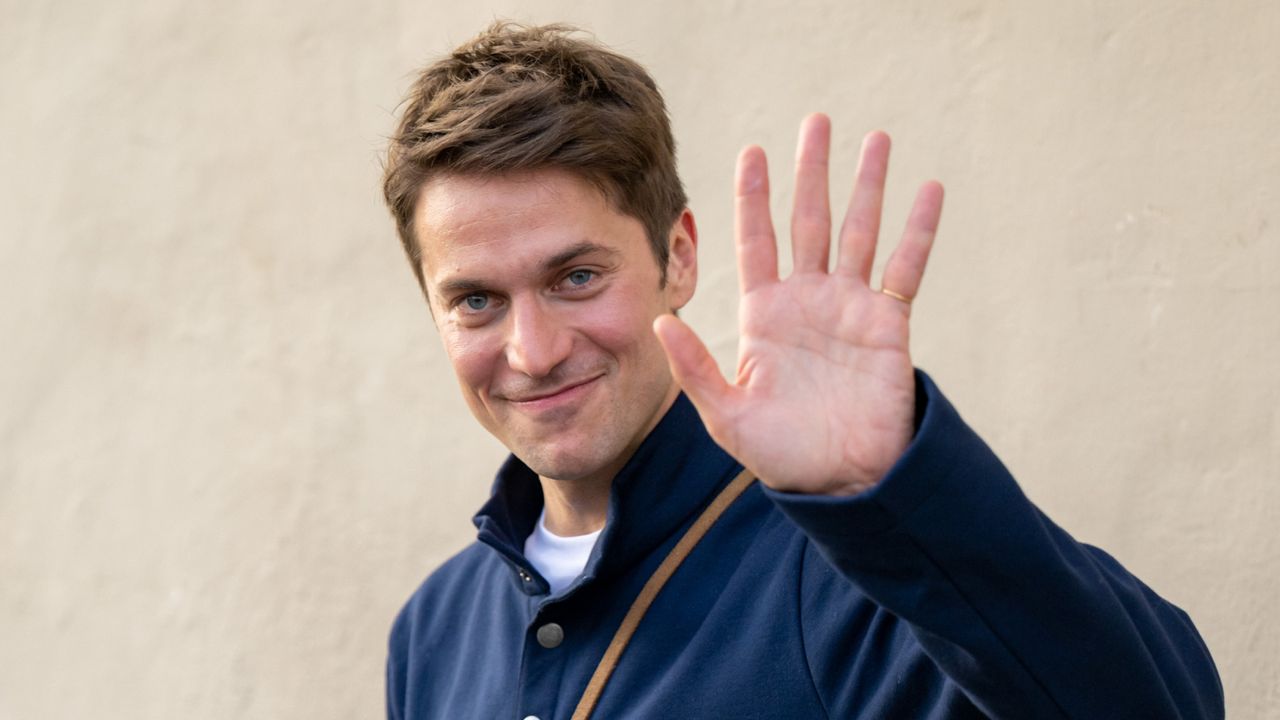 Emily in Paris star Lucas Bravo waving while wearing a navy jacket and carrying a brown leather bag