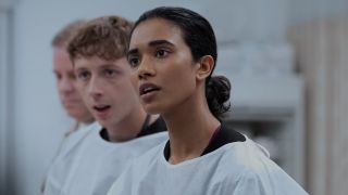 Supriya Ganesh standing in front of Garran Howell on The Pitt. They are both wearing white cover ups over their scrubs. 