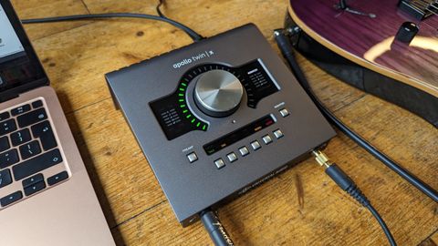 A Universal Audio Apollo Twin X audio interface on a wooden floor with an electric guitar and a MacBook