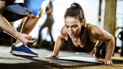 Woman performs push-ups in gym. A man kneels beside her holding a clipboard