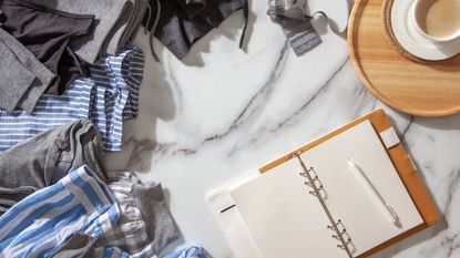 An open planner with a pen next to a cup of coffee, a pile of clothing surrounding it 