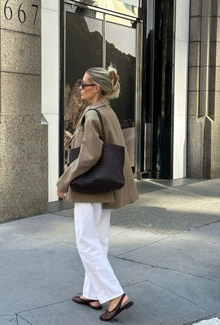 The woven shoe trend is shown in a street-style photo of a woman standing on the sidewalk wearing a tan coat, dark brown tote bag, white jeans, and woven slingback flats