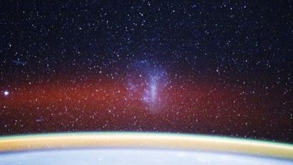 A curved white arc at the bottom of the image glows with a gold band and red fuzzy band of light above it with stars in the distance and a smoky purple shape in the center of the image