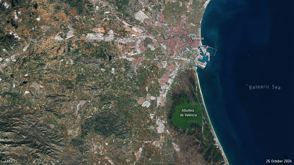 Aerial view from a high altitude, with green populated landmass on the left, and the Balearic Sea on the right. Some small white clouds are sparingly scattered