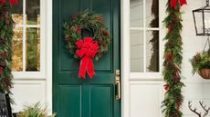 Image of a green front door on a white house with a traditional red and green Christmas wreath hanging on it. There is also a matching garland around the doorway.