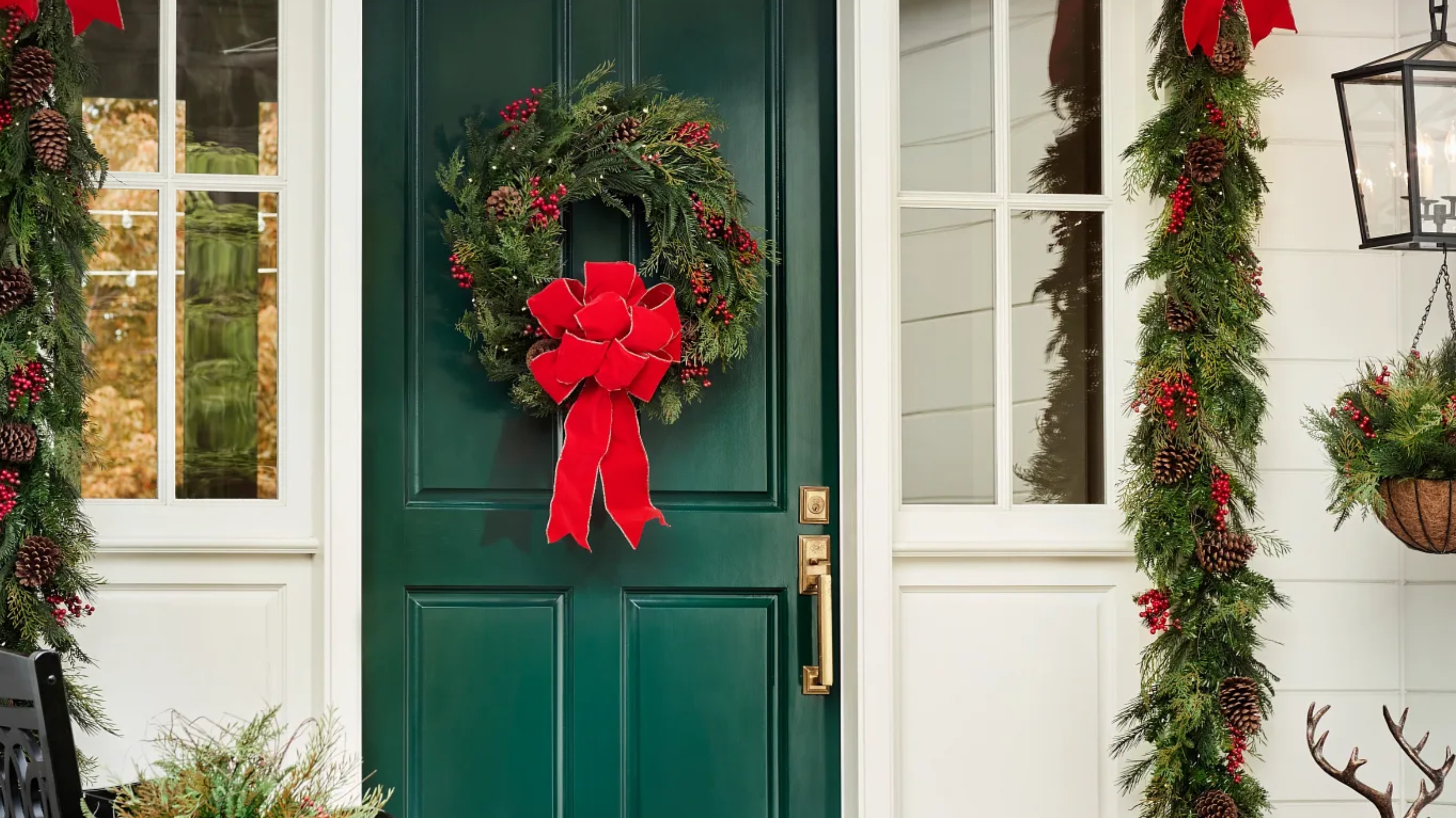 Image of a green front door on a white house with a traditional red and green Christmas wreath hanging on it. There is also a matching garland around the doorway.
