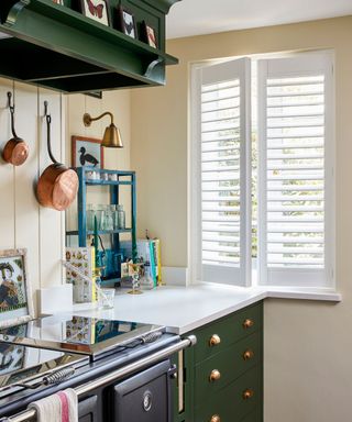 Cream kitchen with green cabinetry, window with white shutters, black Aga, white countertop decorated with kitchenware