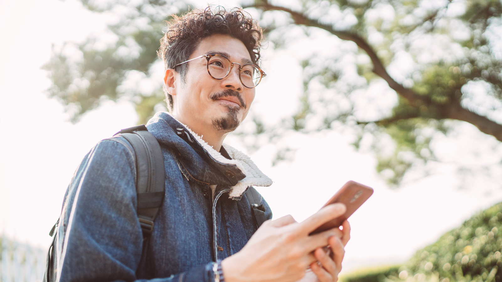 Young man in glasses walking among trees with smartphone in hand, looking ahead
