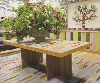 wooden dining table with colourful mosaic top on colourful checked rug in colourful space with tree mural and large vase of foliage on top