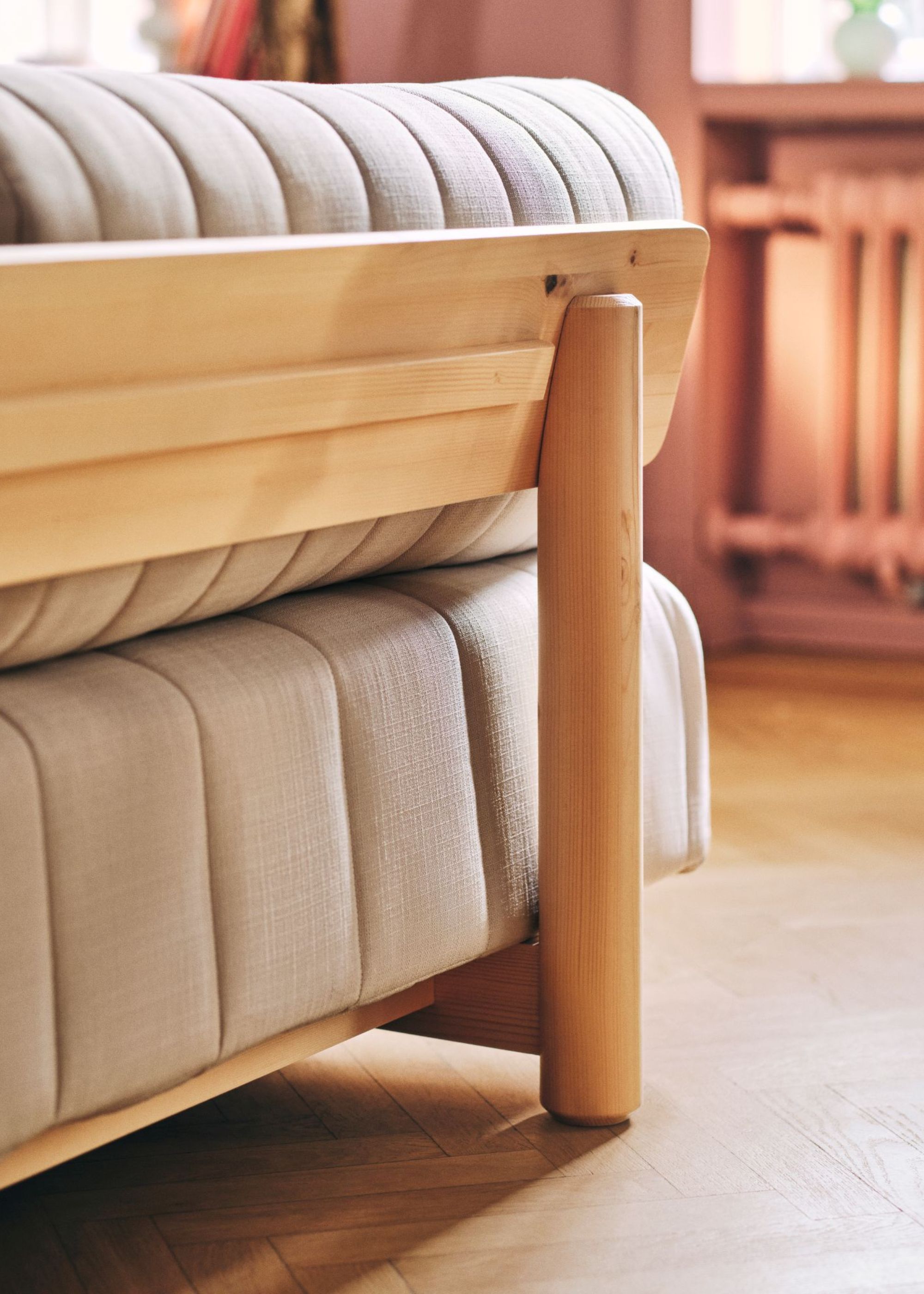 The back corner of a cream-gray sofa bed with a wooden base and frame on a wooden floor in a living room with the radiator just slightly in view in the background