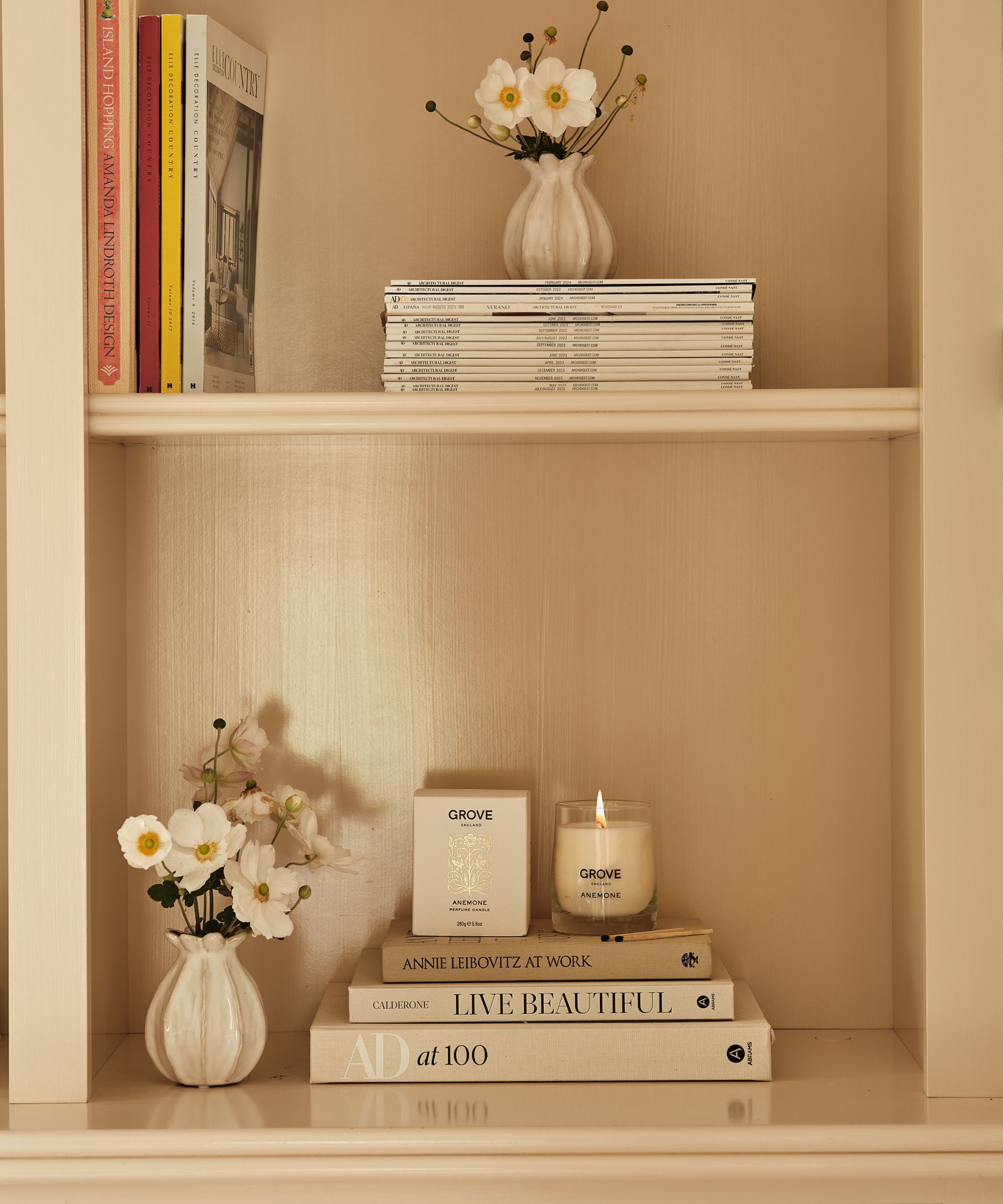 Candle, books, magazines and flower jars on a cream bookshelf