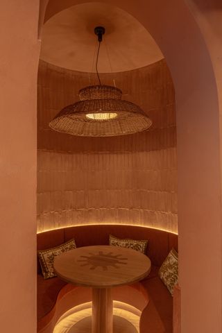 The interiors of a modernly furnished restaurant feature woven rattan lamps, suffused lighting, vibrantly decorated banquettes, and terracotta brick walls.