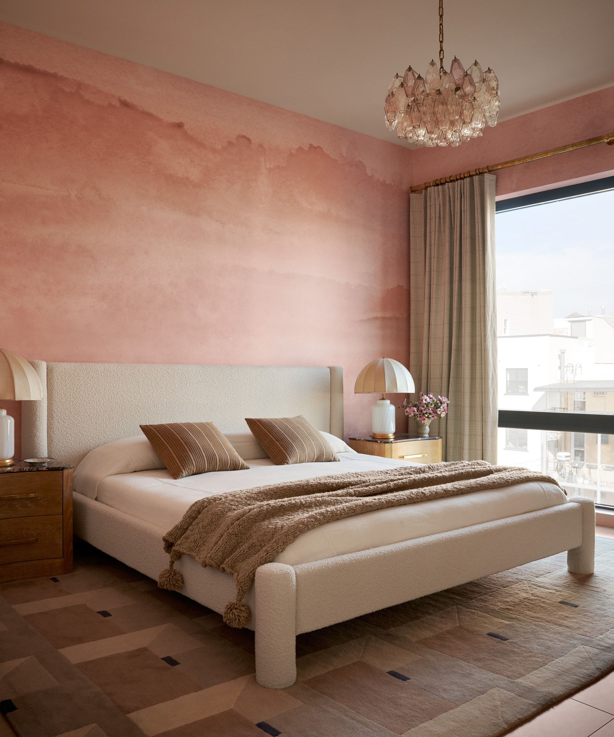 Pink bedroom with cloud mural, chandelier, large boucle bed frame and neutral bedding and nightstand with lamp and vase