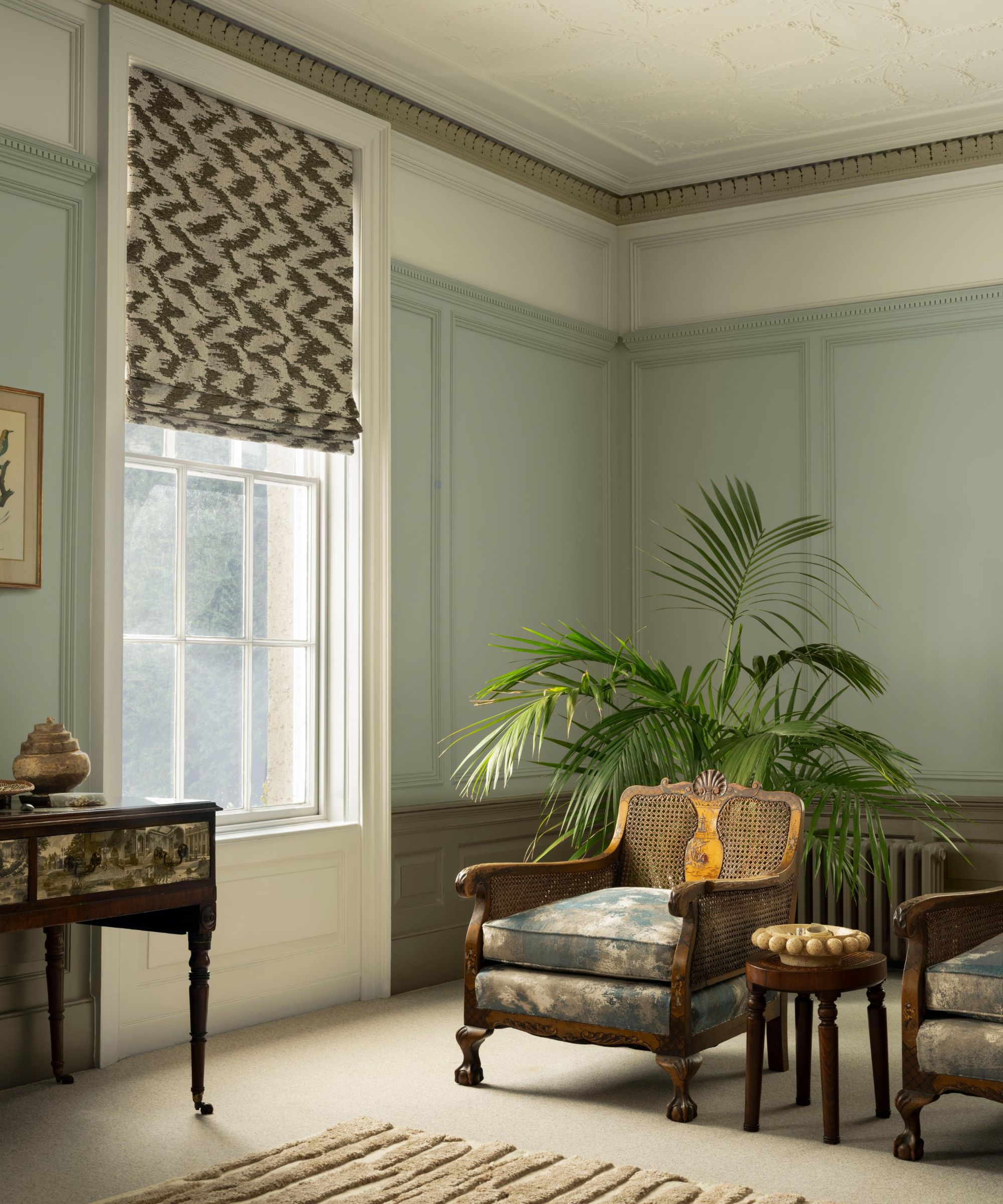 Living room with pale blue walls, cream carpet, vintage mahogany cane armchair and side table, a textured beige rug, and a large window.