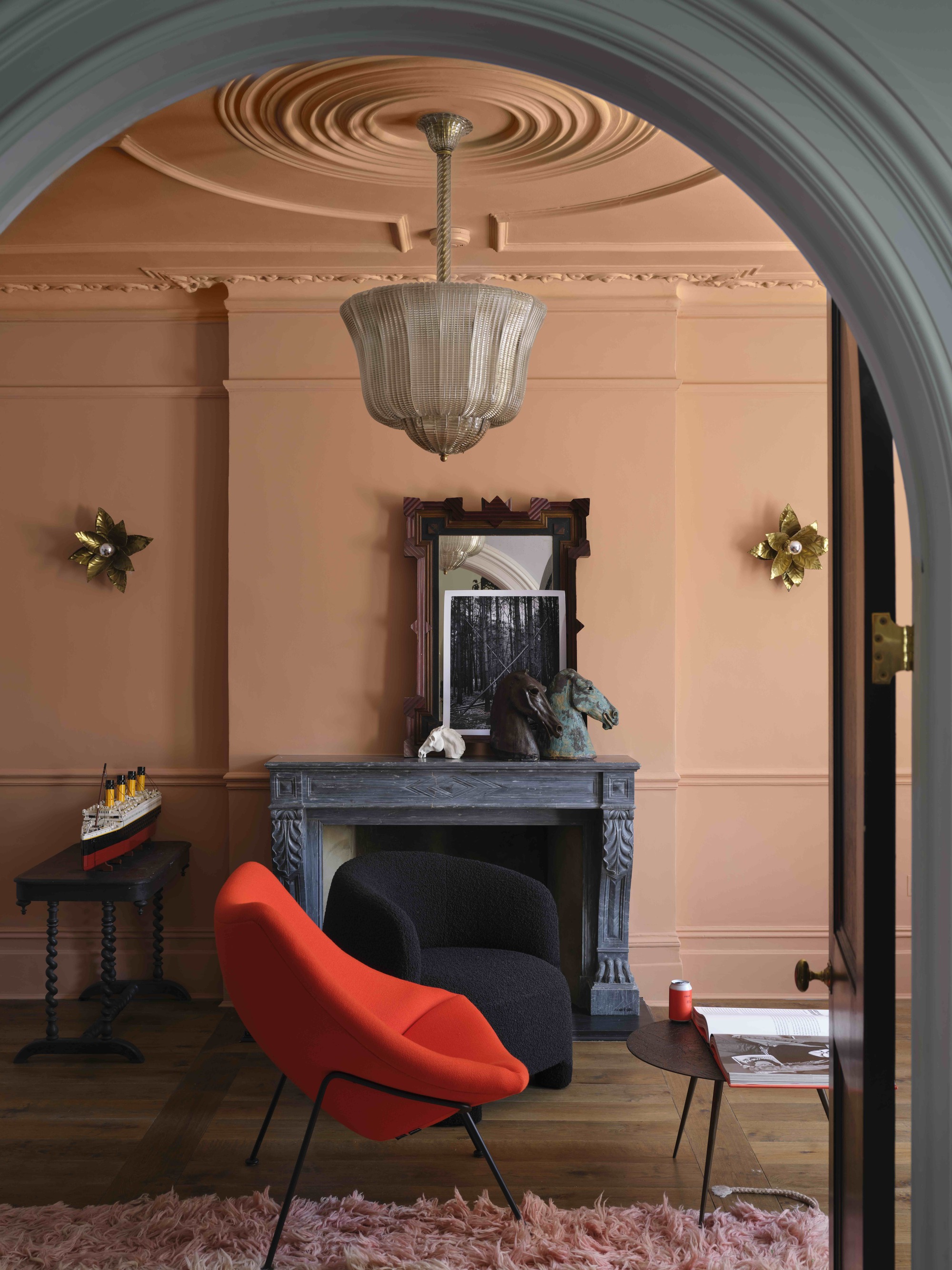 a color drenched peach room with decorative ceiling and wall molding, an ornate dark fireplace with a mirror and picture styled on the mantel, a mesh pendant light, and gold sconces on the wall, a fluffy pink rug, and red and black armchairs in front of a coffee table
