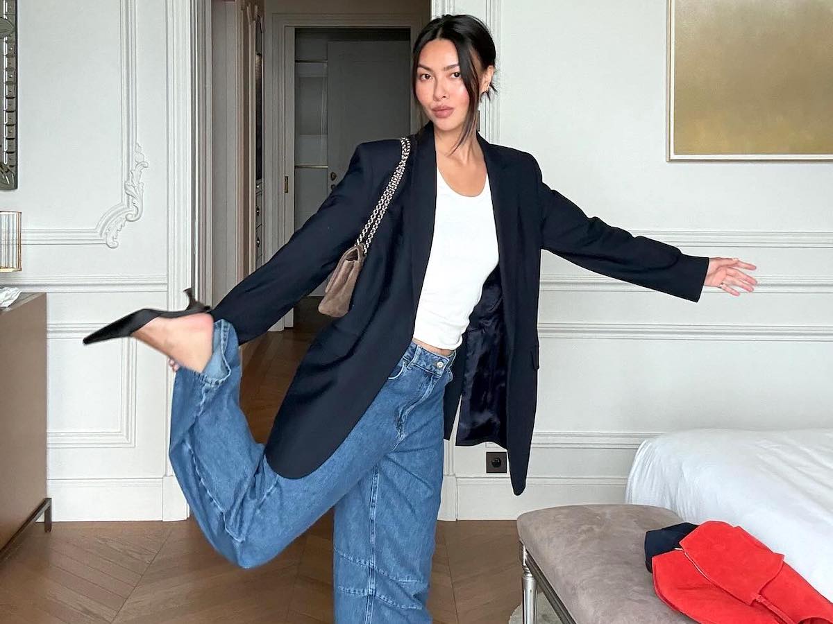 Mimi Nguyen poses with one leg up in a chic hotel in Monaco wearing a black blazer, white t-shirt, a taupe chain-strap bag, baggy barrel jeans, and black mule heels