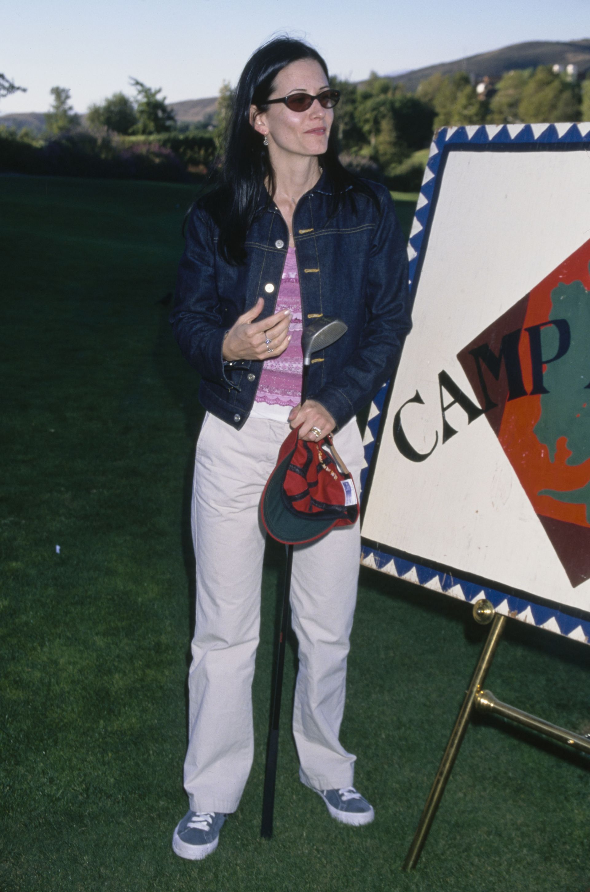 Courtney Cox in 1999 wearing dark grey trainers with a white trousers, a pink striped tank top and an indigo denim jacket.