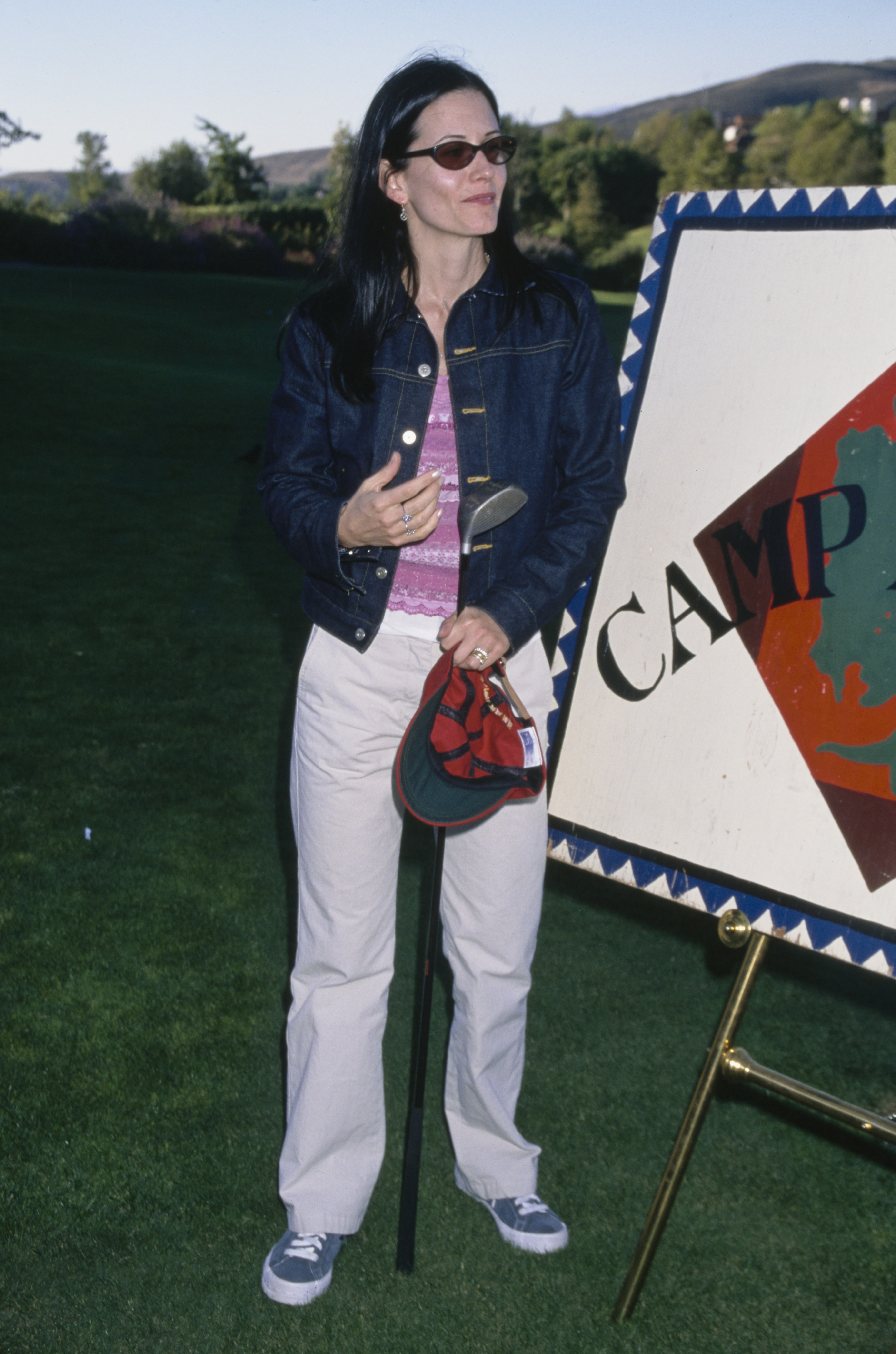 Courtney Cox in 1999 wearing dark grey trainers with a white trousers, a pink striped tank top and an indigo denim jacket.
