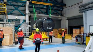 a cross-bar crane hoist hanging from a support beam across the ceiling of a warehouse suspends a large black cylinder with two green straps, connected to the cylinder at four points. Four workers in orange highlighter apparel stand around the suspended cylinder, and another stands off to the left.