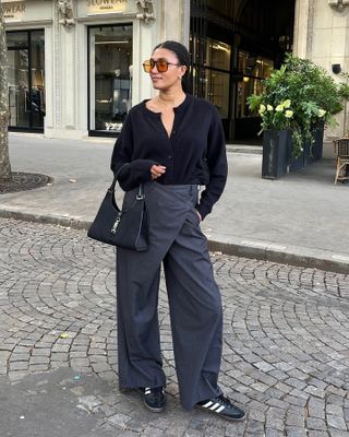 French influencer Lena Farl poses on a cobblestone street in Paris wearing orange tinted oversize sunglasses, a black cardigan, a black shoulder bag, gray wrap pants, and black Samba sneakers.