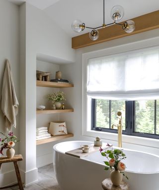 A white bathroom with wood ceiling beams and matching wood shelving in an alcove. A freestanding white bathtub sits under the window with a wooden bath caddy topped with a decor. A god tap sits by the bath and a brass chandelier hangs above the bath