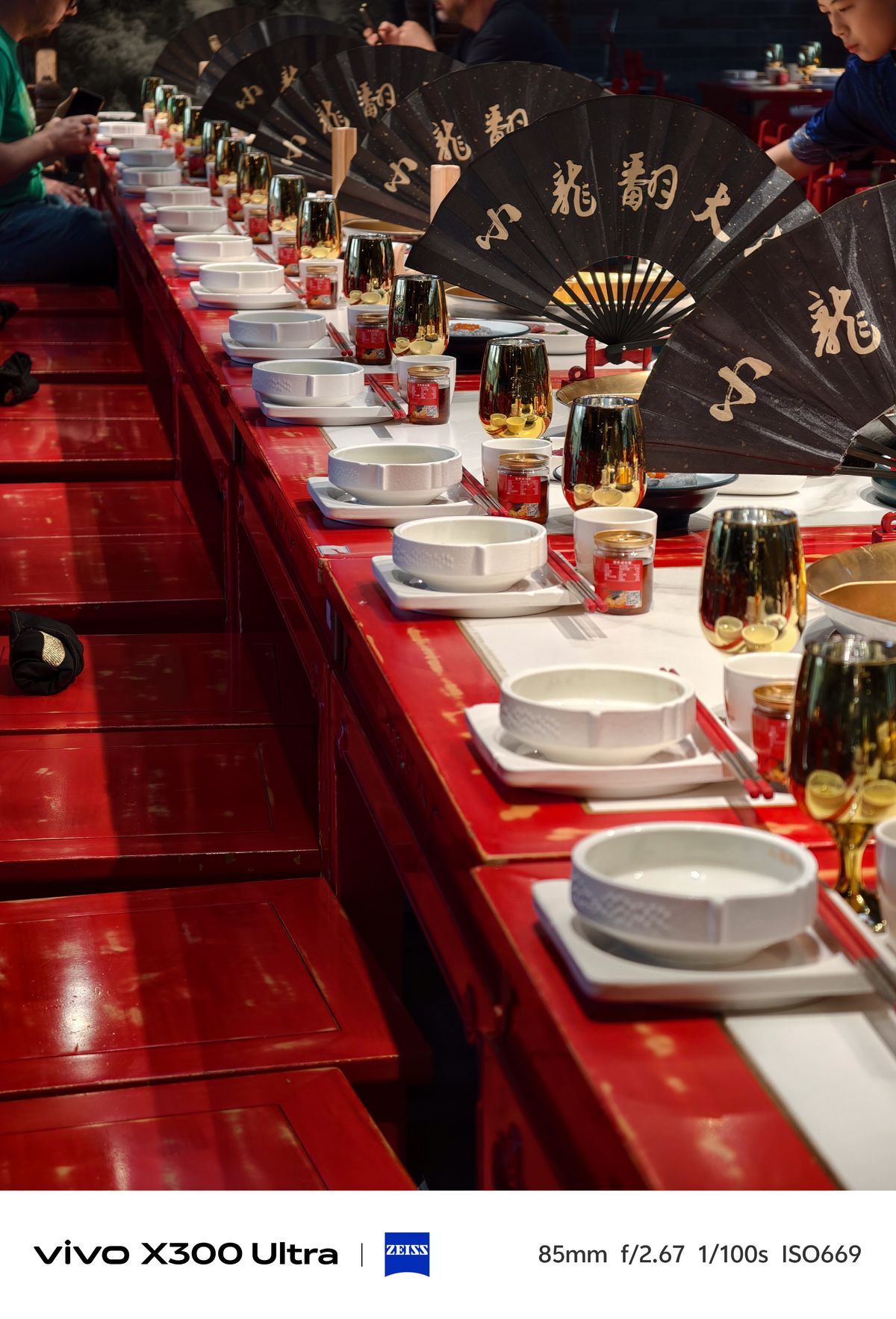 Long banquet table set with white bowls, gold cups, and decorative black paper fans.