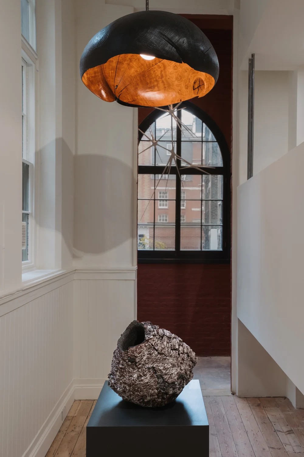 A hallway filled with a raw, vase-like sculpture placed atop a wooden black podium, topped by a wooden lamp shade with an organic shaped.
