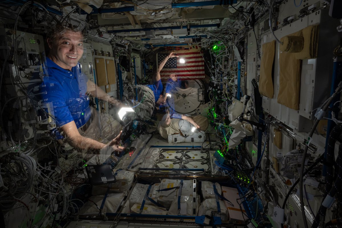 Una pila de fotografías de Matthew Dominick que muestran varias imágenes fantasmales de sí mismo en un módulo oscuro de la estación espacial.  detrás hay una bandera americana