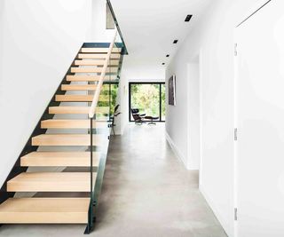 A white hallway in a contemporary home. The hallway features concrete-effect flooring, a large picture window to one end, which frames the view of the garden, and a modern timber and steel open tread staircase from Complete Stair Systems.