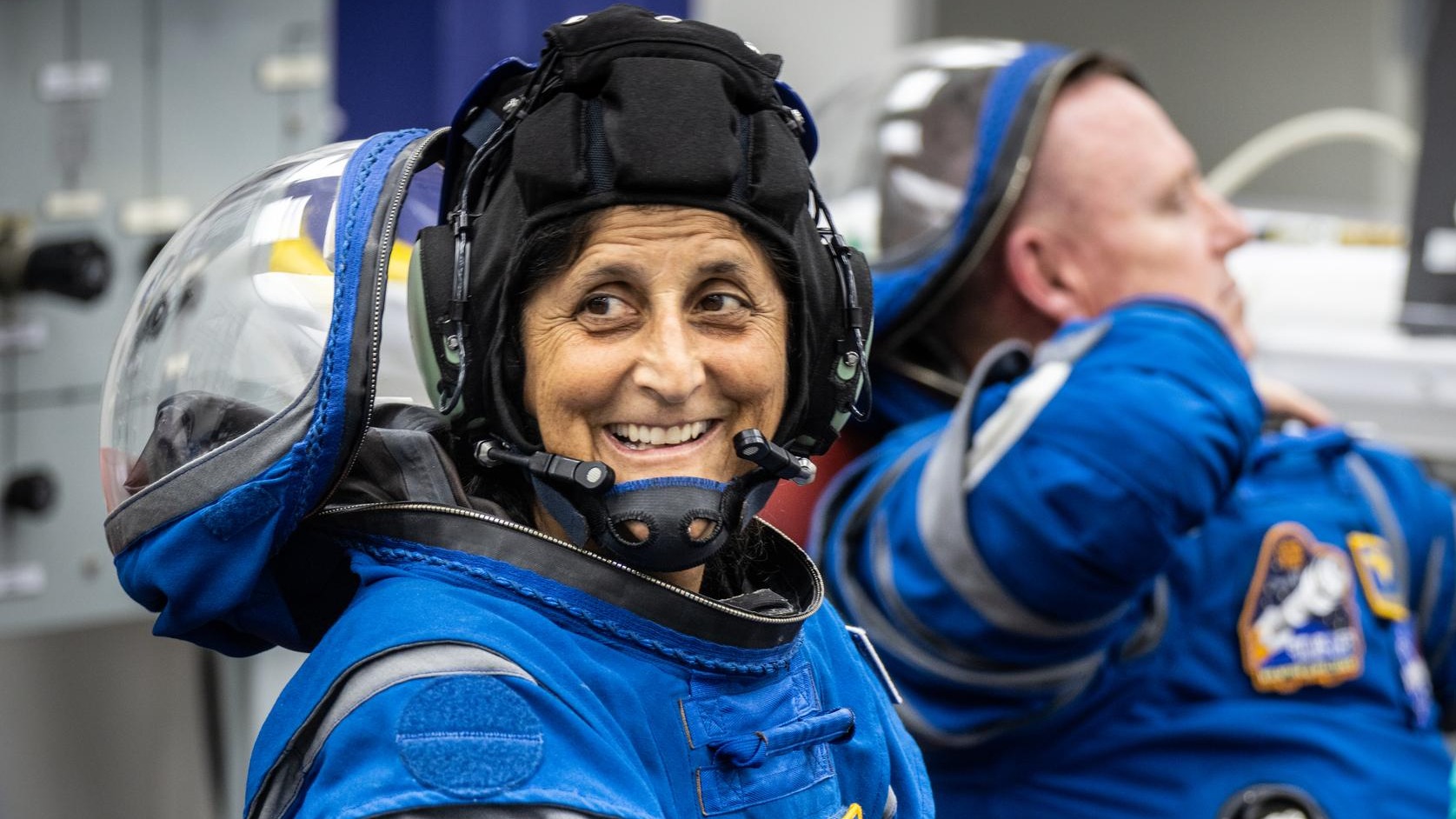 Two astronauts in space suits with visors open: Suni Williams in front, smiling at the camera, and Butch Wilmore in the back, looking off-screen.