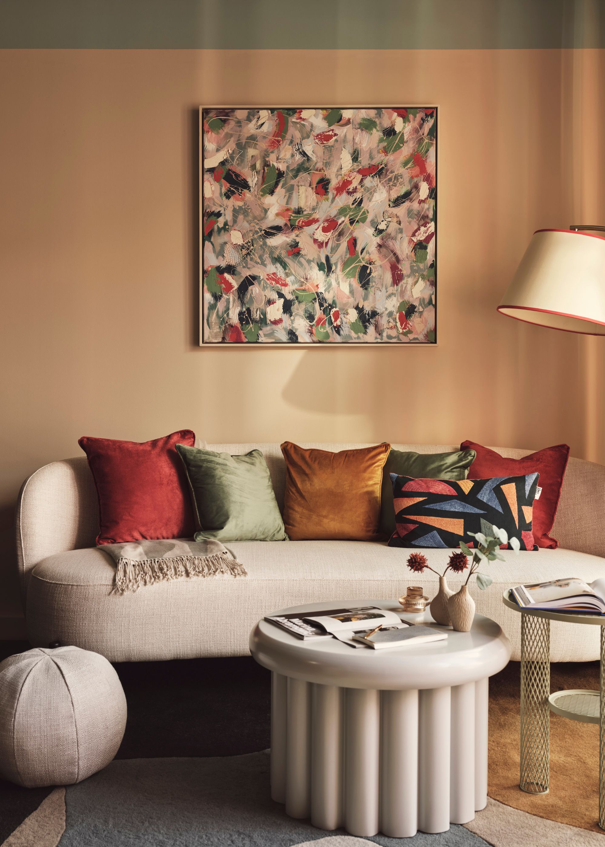 A modern living room with peach walls, abstract artwork on the wall, a taupe curved sofa with colorful cushions, and a modern round coffee table.