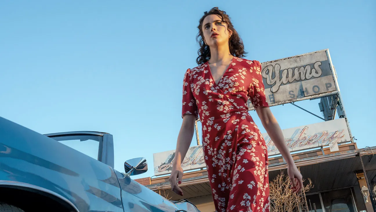 margaret qualley wears a red floral dress walking by a car in a still from the movie honey don't