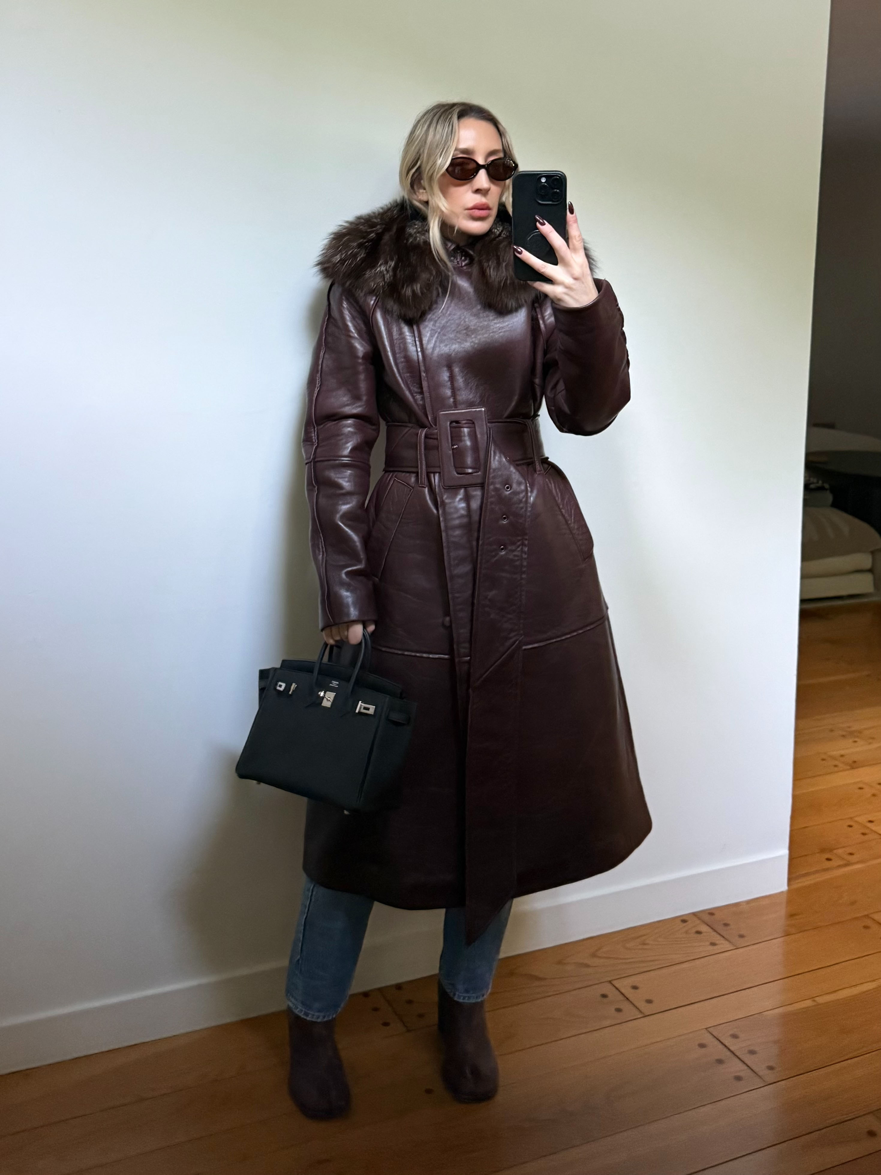 Woman taking a selfie in a brown collared jacket, oval sunglasses, black Birkin handbag, and wavy blonde hair holding an iPhone