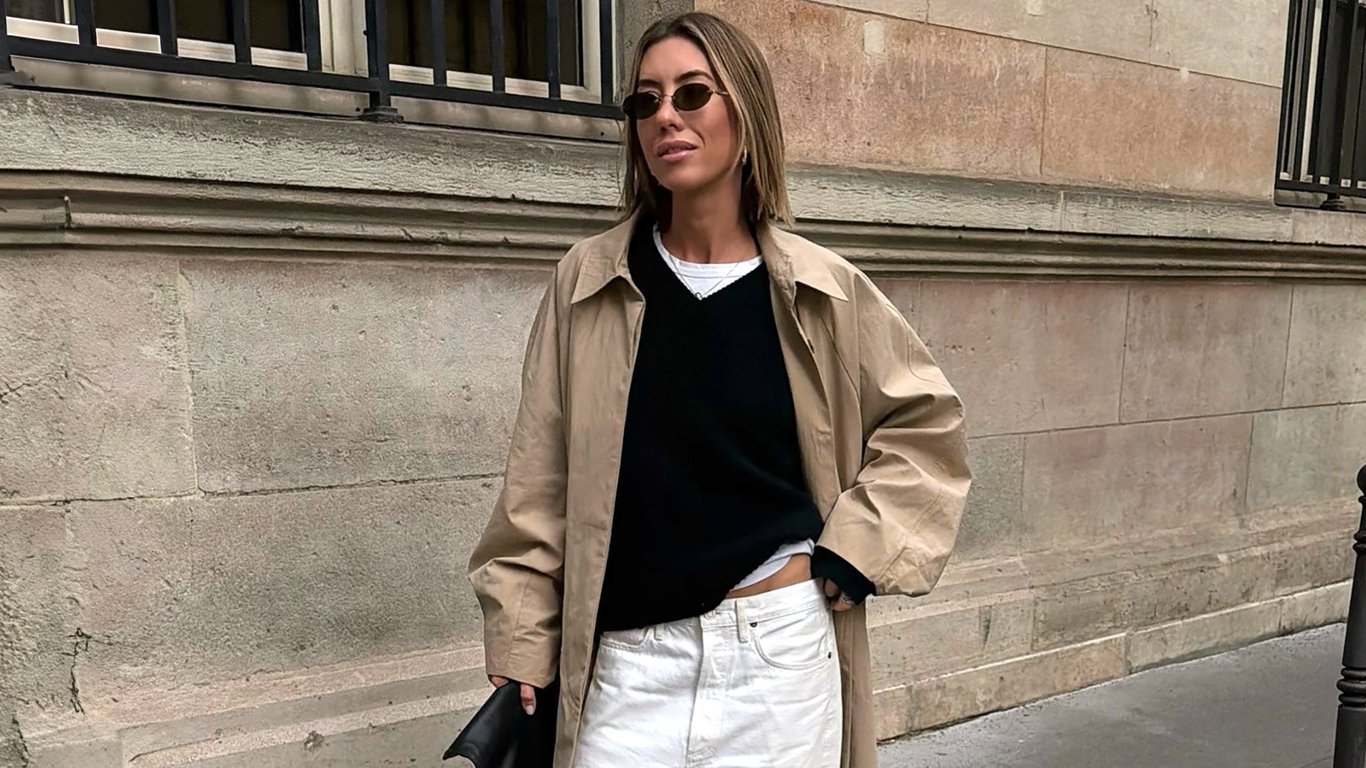 stylist and fashion influencer Hannah Lewis poses on a sidewalk in the UK wearing small oval sunglasses, a trench coat, a white T-shirt, black V-neck sweater, and white jeans
