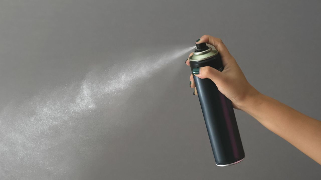 A woman&#039;s hand spraying a can of dry shampoo on a grey backdrop