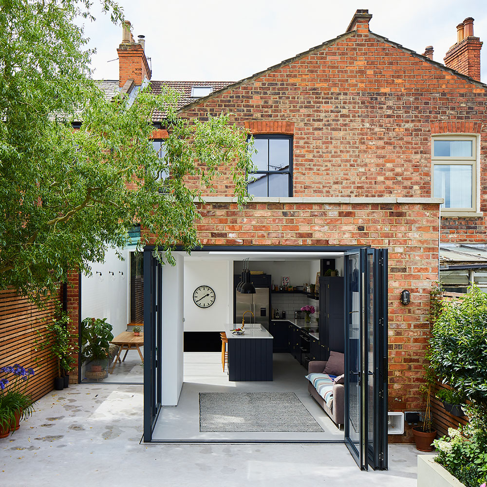 Rear view of a brick house with an extension with full width bi-fold doors which are open revealing a kitchen.