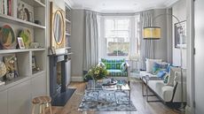 Modern living room with grey walls, white furniture and a glass coffee table on a blue rug. Above a dark blue painted mantelpiece is a gold mirror, and to the left are shelves with colorful plates, books and vases.