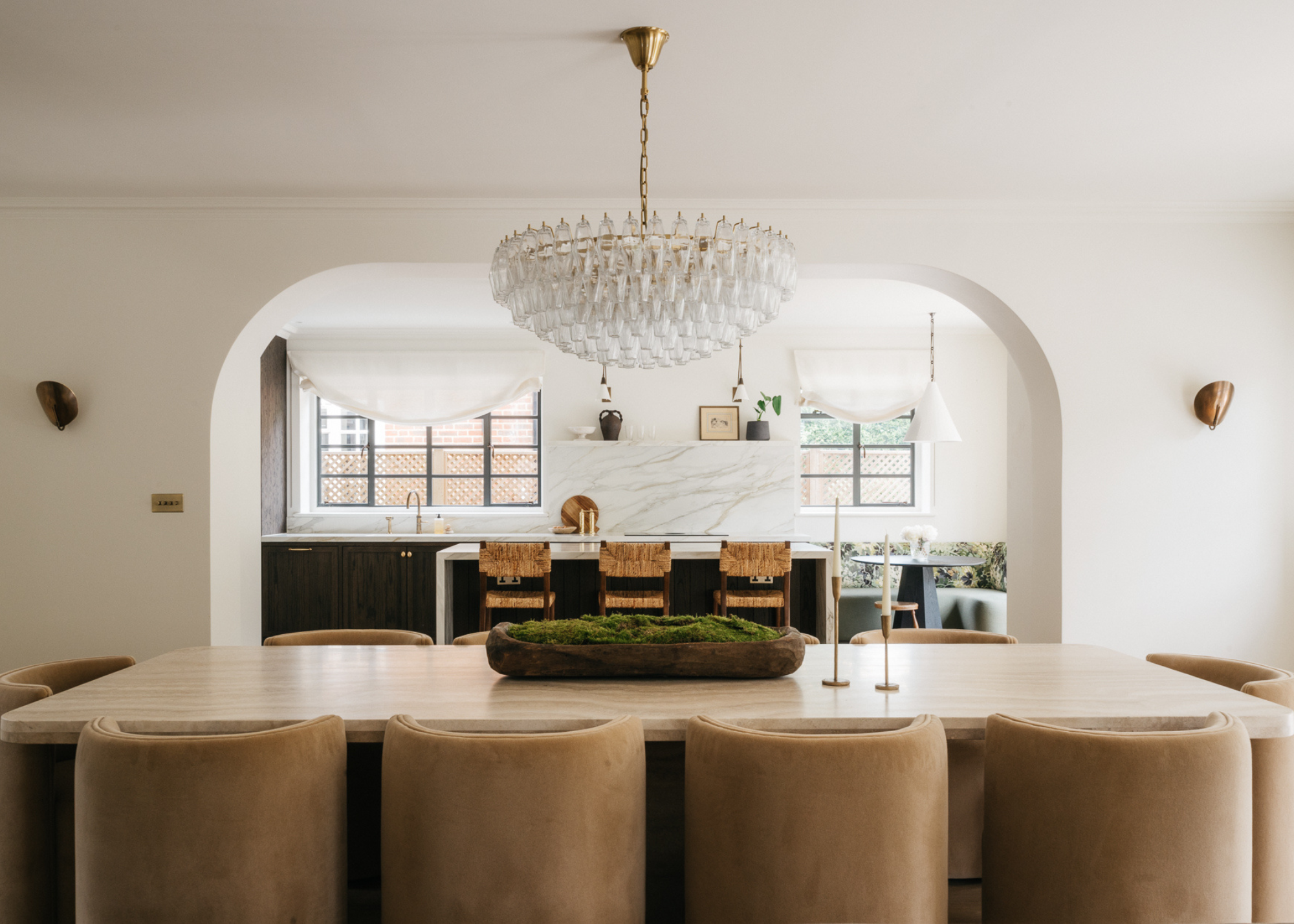 A large dining room table with curved armchairs in a neutral space with an archway leading through to a modern kitchen with a kitchen island and bar stools and banquette seating in view in the corner