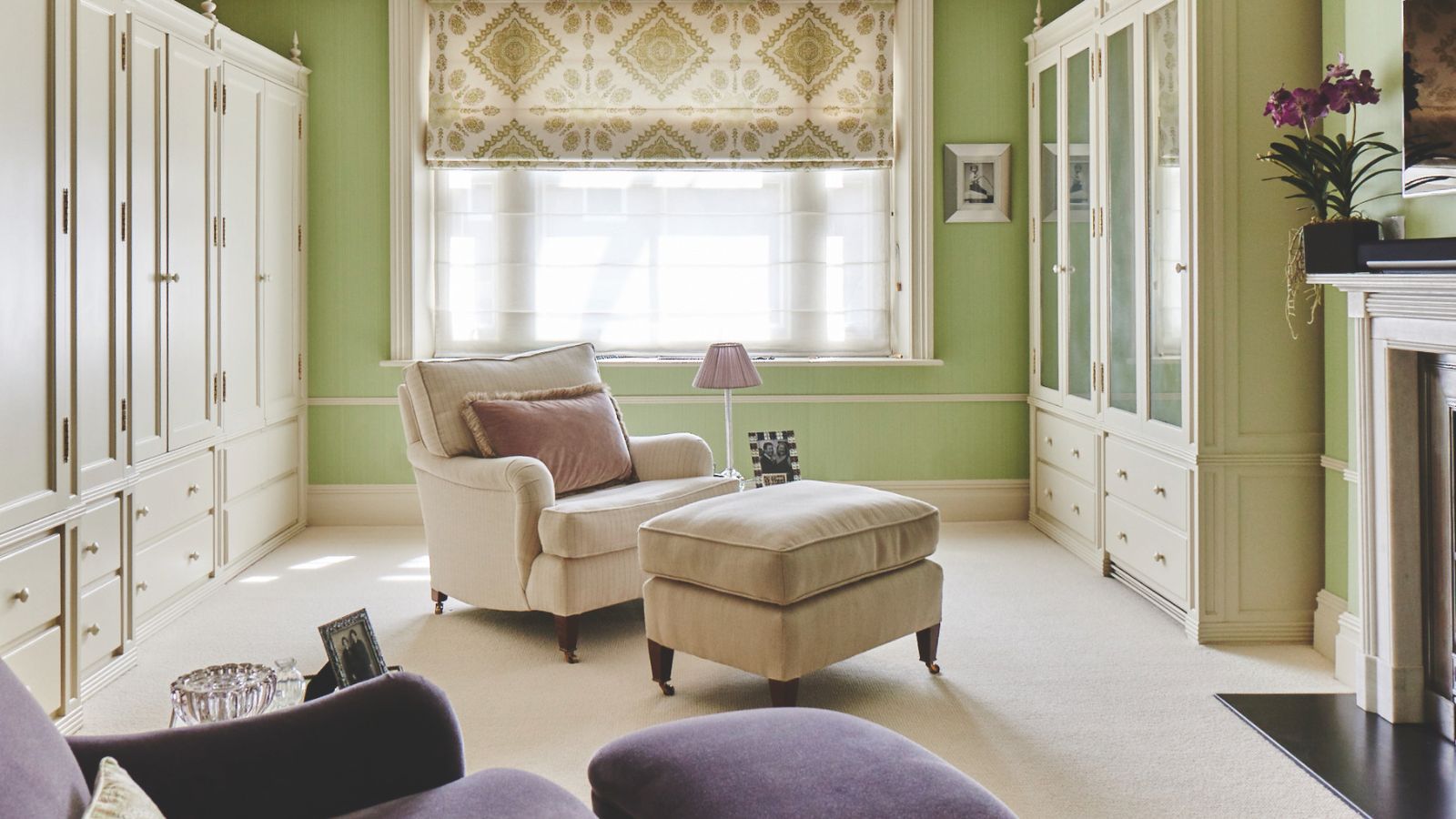 A green dressing room with white carpet, white built-in wardrobes, a large window with a sheer curtain and green and white patterned blind, and a white fireplace to the right. In the center of the room is a white upholstered chair with matching footrest and pink lamp and cushion, and in the foreground is a purple upholstered chair.