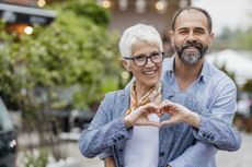 Cheerful mature couple is embracing on the street, they are looking at the camera and woman is making hear shape symbol with her hands