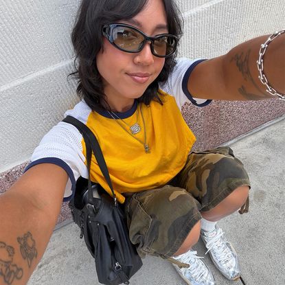 @shhtephs wearing a Baseball tee, camo shorts, and silver sneakers