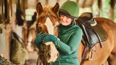 A woman wearing a green baselayer and riding helmet with a green silk cuddles the head of a chestnut horse, who is wearing. saddle and bridle with a sheepskin noseband
