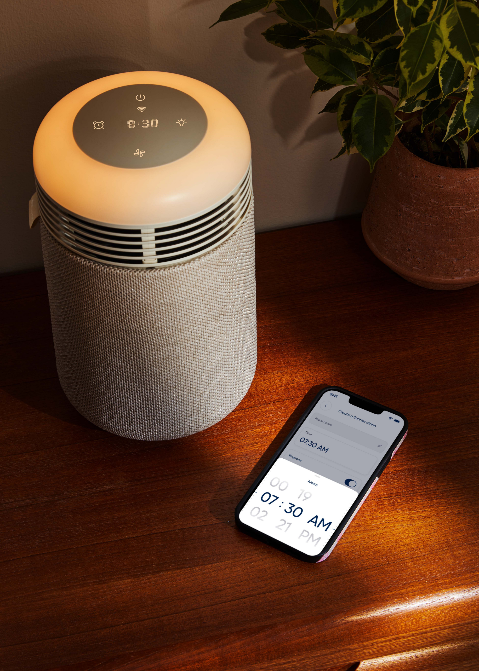 A gray air purifying sun clock by a phone with an alarm and a potted plant on a wood side table