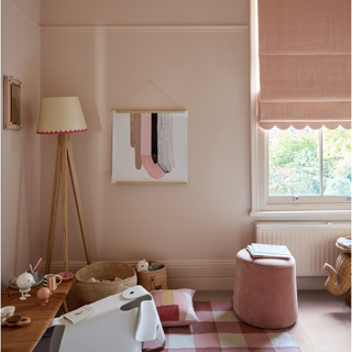 A pink painted childrens room with pink checked rug and scalloped edge blind over window