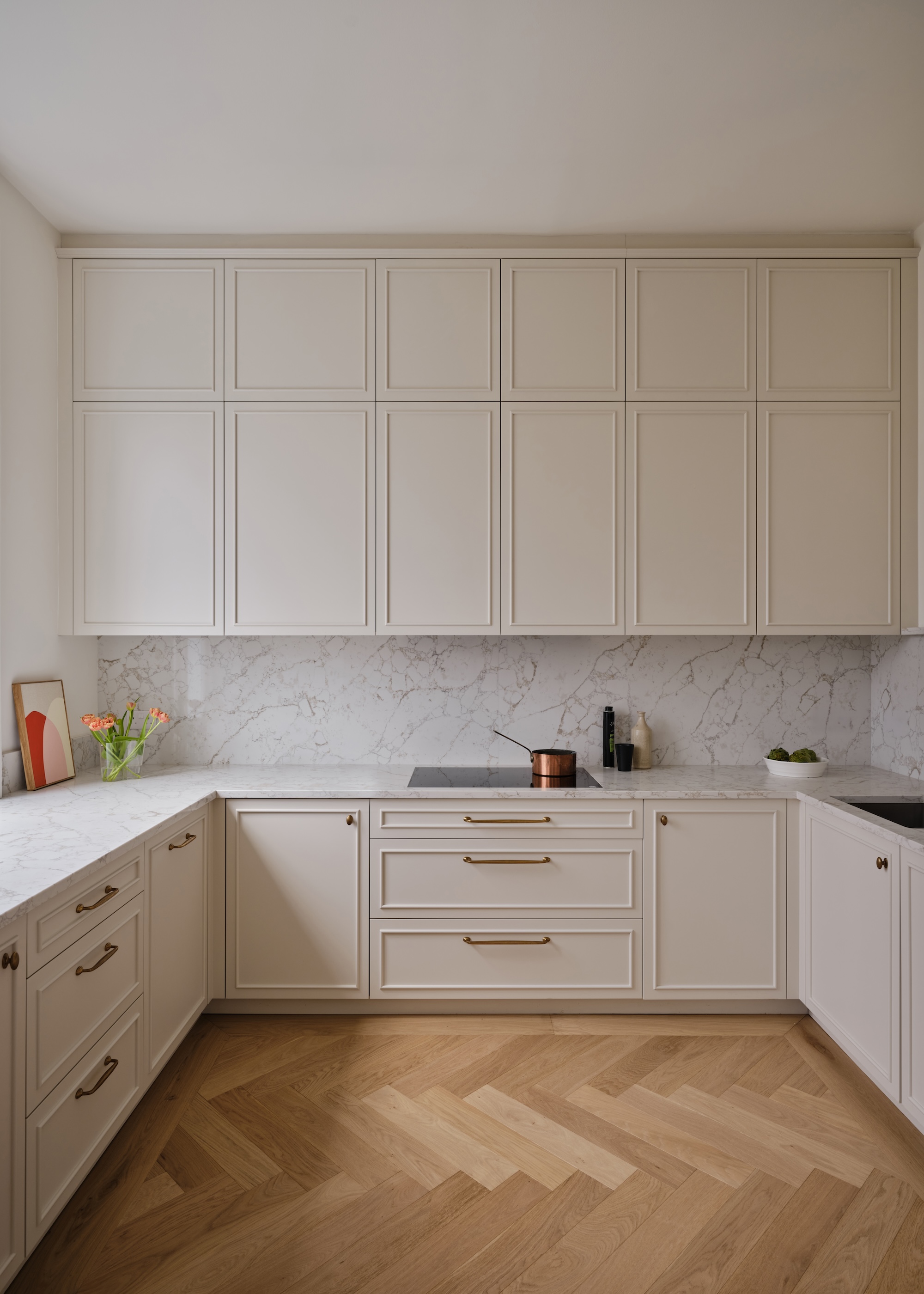 a classic Shaker kitchen in off white showing handleless upper wall units and with handles on the base cabinets, plus herringbone wood flooring and marble worktops and splashback