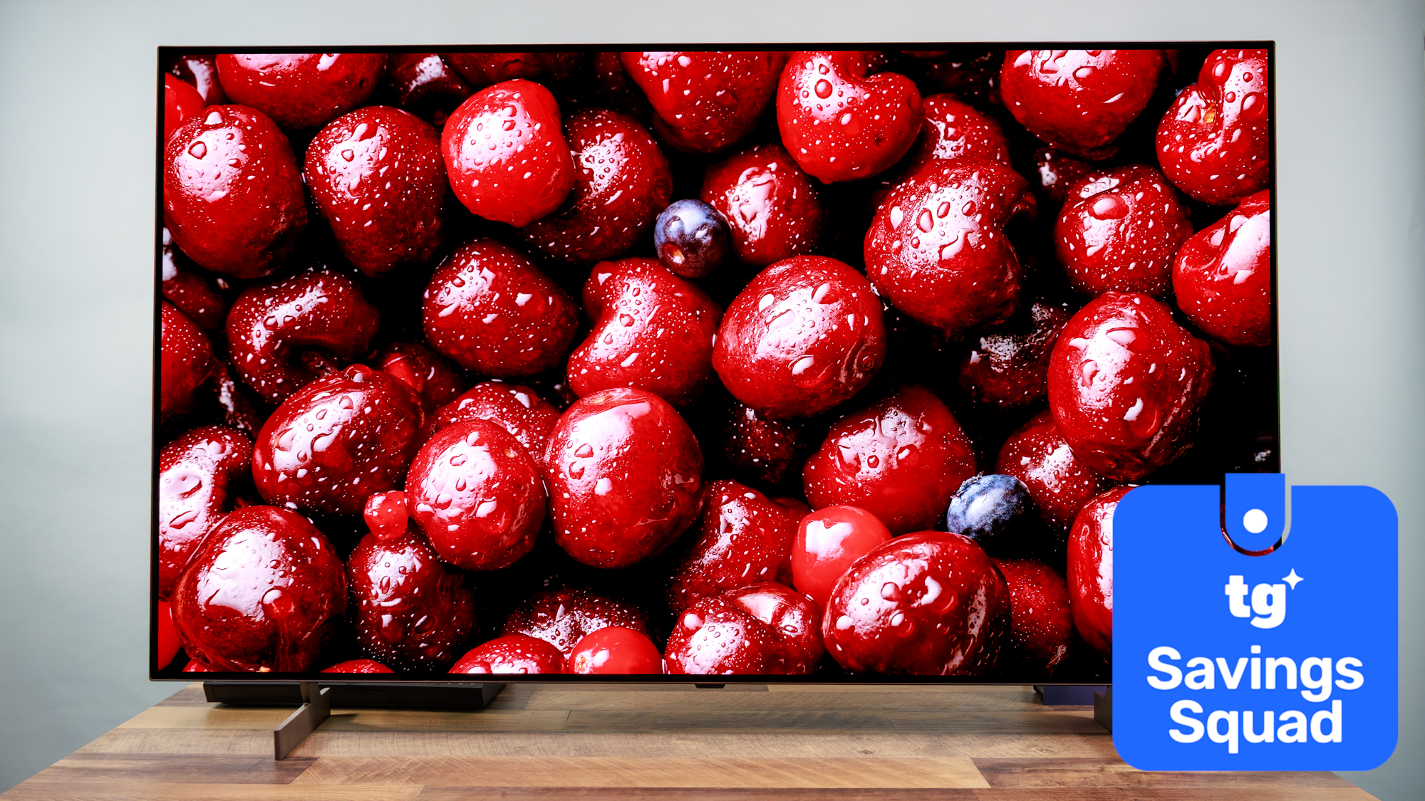 The LG B6 OLED TV displaying a high-contrast, close-up image of dark red-colored cherries. Next to the TV is a superimposed badge that reads: "TG Savings Squad".