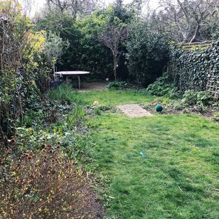 A poorly-maintained garden with uneven lawn and grey outdoor table at rear of garden