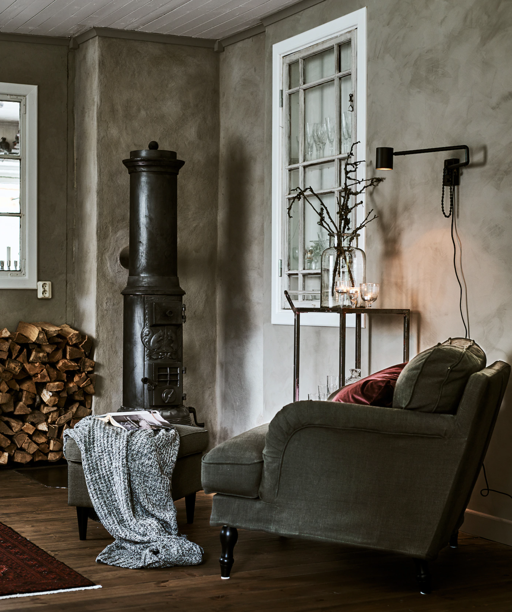 A grey living room or snug with black fireplace, grey armchair, concrete walls and logs in corner for burning
