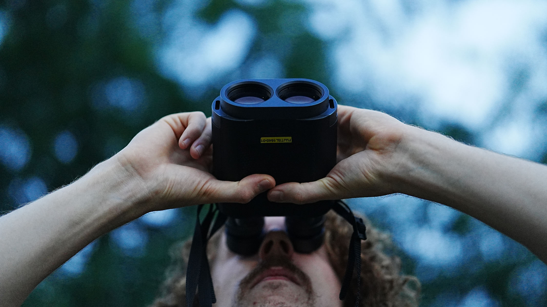 A man looking through the Fujifilm Techno-Stabi TS-L 1640 image-stabilized binoculars upwards with trees in the background.